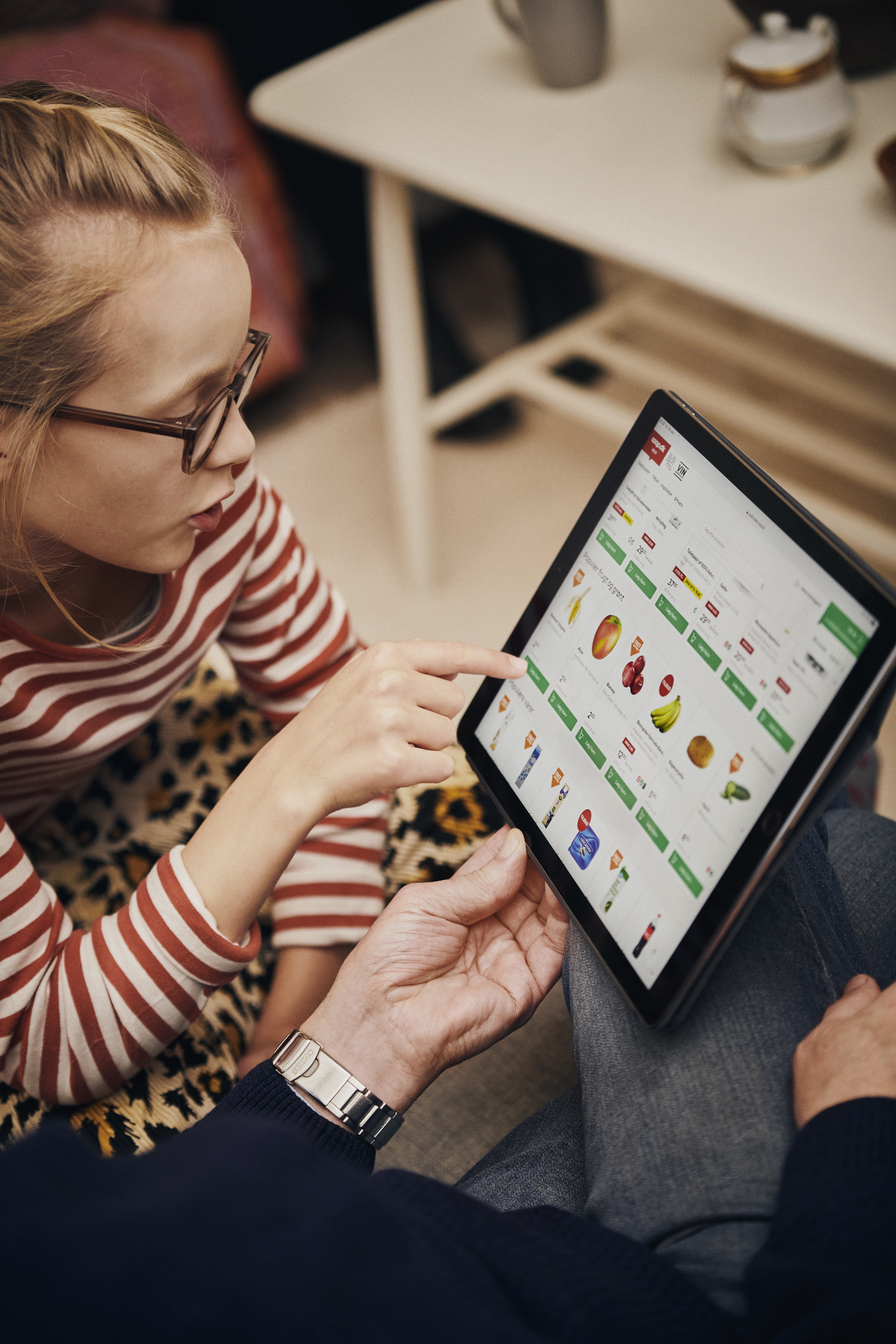 A child in striped clothing points at a digital grocery shopping interface on a tablet while an adult's hand is visible holding the device, showcasing a variety of food items for purchase.