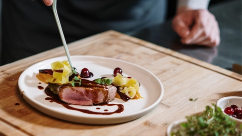 En kok anretter en tallerken med saftig and, farverig grøntsags garniture og glaserede kirsebær på et træskærebræt med en moderne, stilren indretning.