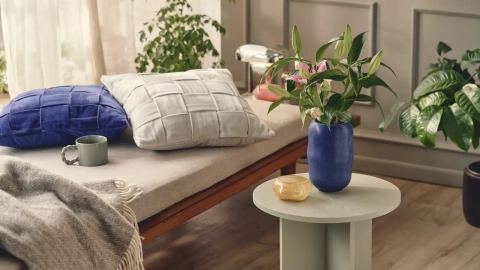 Lys stueindretning med en sofa dækket af en grå plaid, to puder i blå og grå farver, en grøn kop, en vase med blomster og en lille sidebord i lys farve. Planteliv i baggrunden.