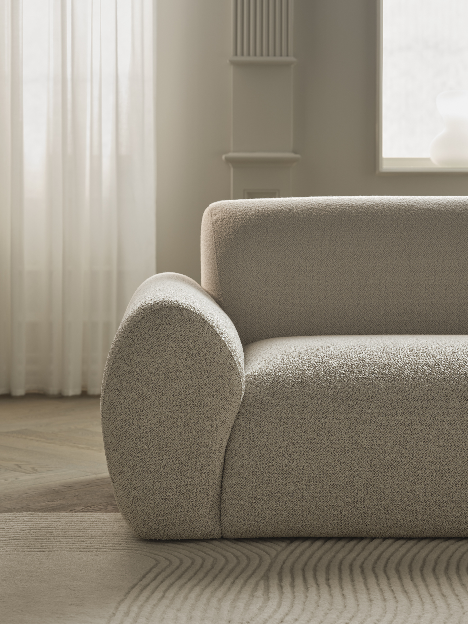 En moderne sofa med bløde kurver i en lys beige farve, placeret på et mønstrede tæppe, med lys, der filtreres gennem en hvid gardin i et stilrent rum.