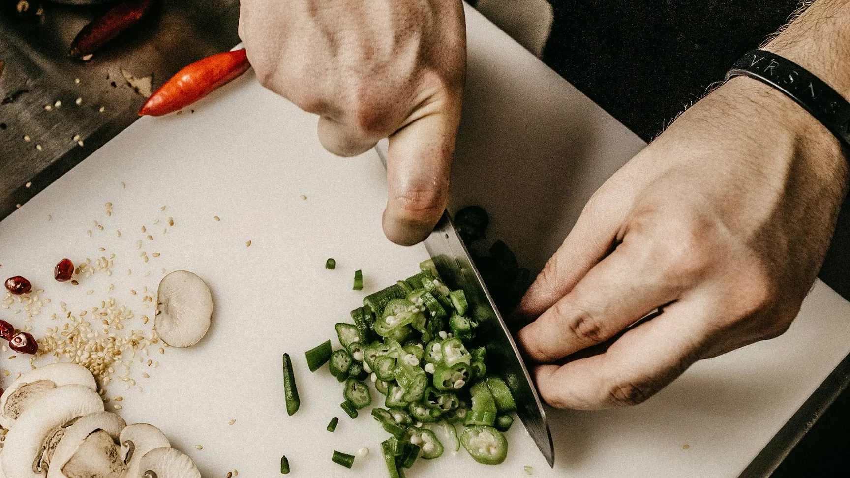 En kok skærer grønne løg på et skærebræt i et moderne køkken, omringet af forskellige ingredienser som svampe, sesamfrø og røde chili.