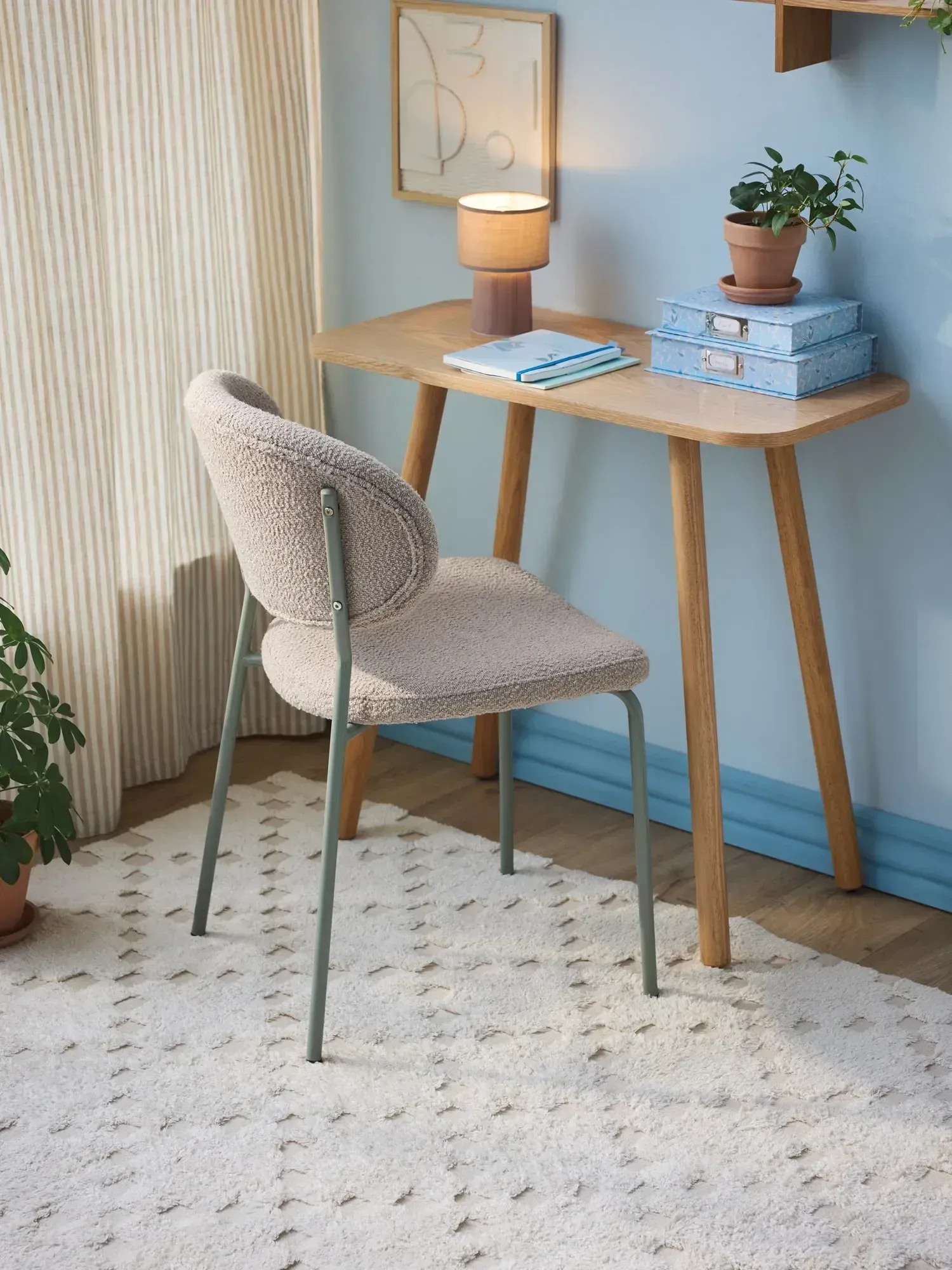 En moderne stol i beige stof står ved et træbord med en lampe, bøger og en potteplante, beliggende på et mønstret tæppe i et lyst rum.