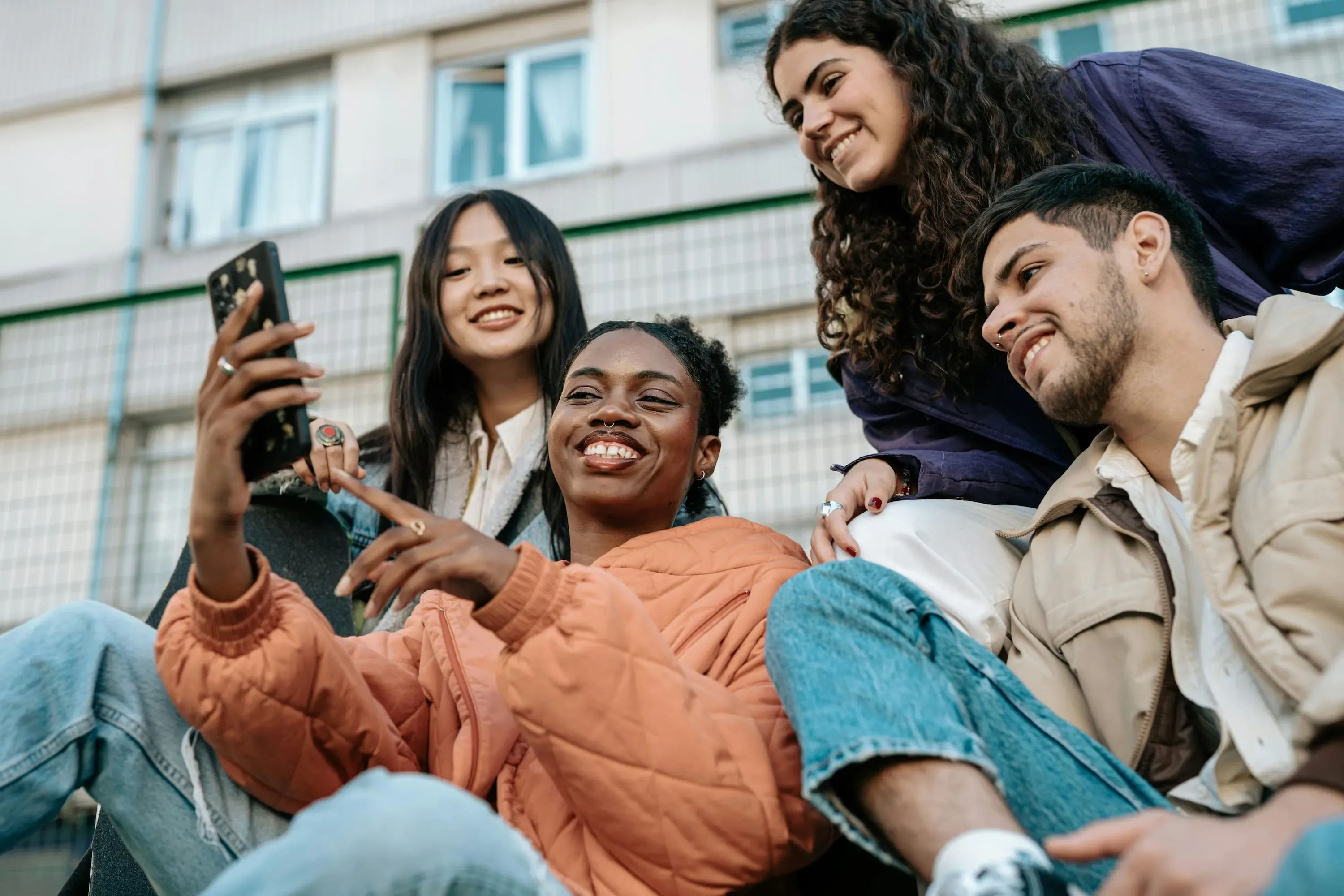 Fire unge voksne smiler og kigger på en smartphone sammen, mens de sidder på en trappe. De har forskellige hårtyper og stilarter, iført afslappet tøj. I baggrunden ses en bygning med vinduer og et gitter.