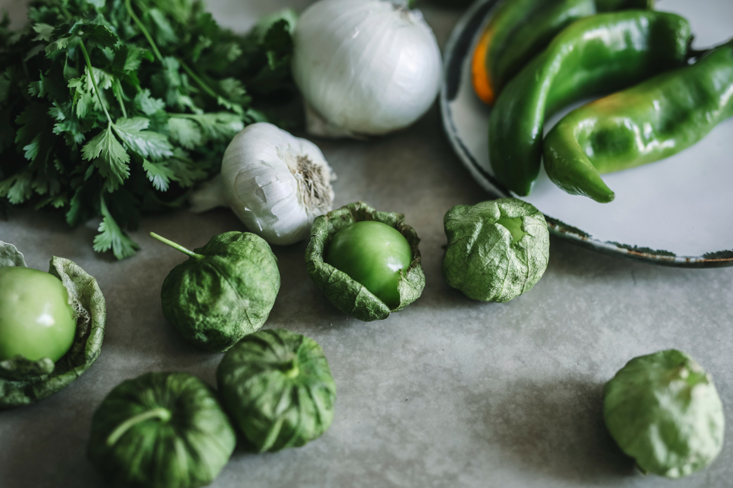 Friske grønne grøntsager herunder koriander, hvidløg, tomatillos og grønne chili på en bordplade.