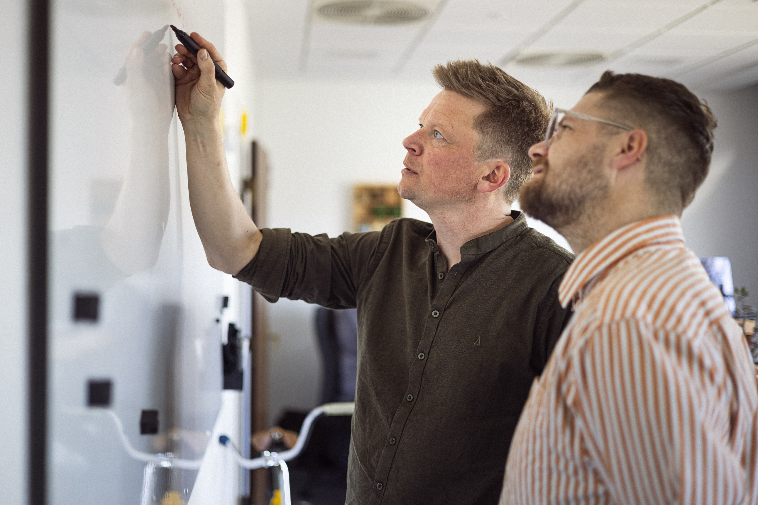 To personer i et kontormiljø står ved et whiteboard, hvor den ene person skriver med en tusch, mens den anden ser opmærksomt på.