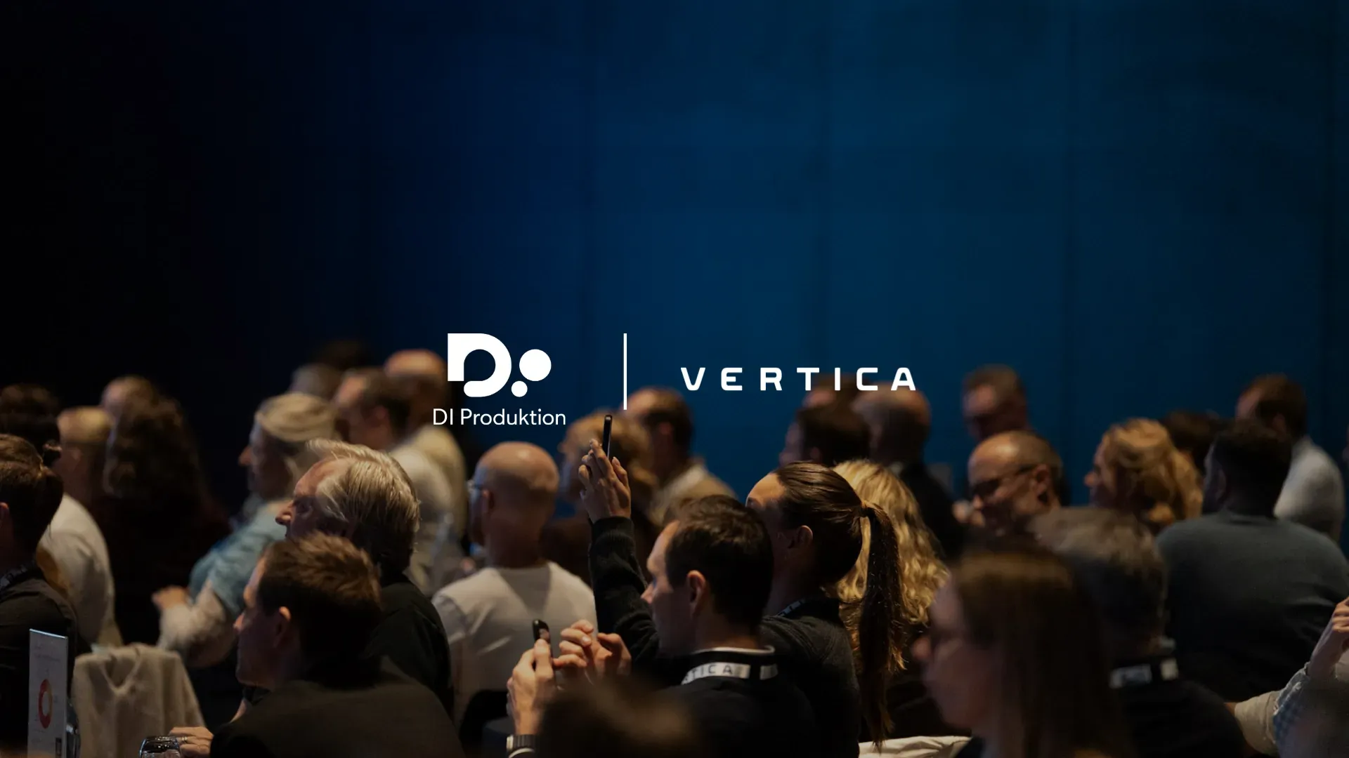Billede af en stor forsamling, der deltager i et arrangement, hvor folk sidder og kigger mod scenen, med fokus på det kollektive engagement i en professionel konference. Logos for DI Produktion og Vertica vises i midten.