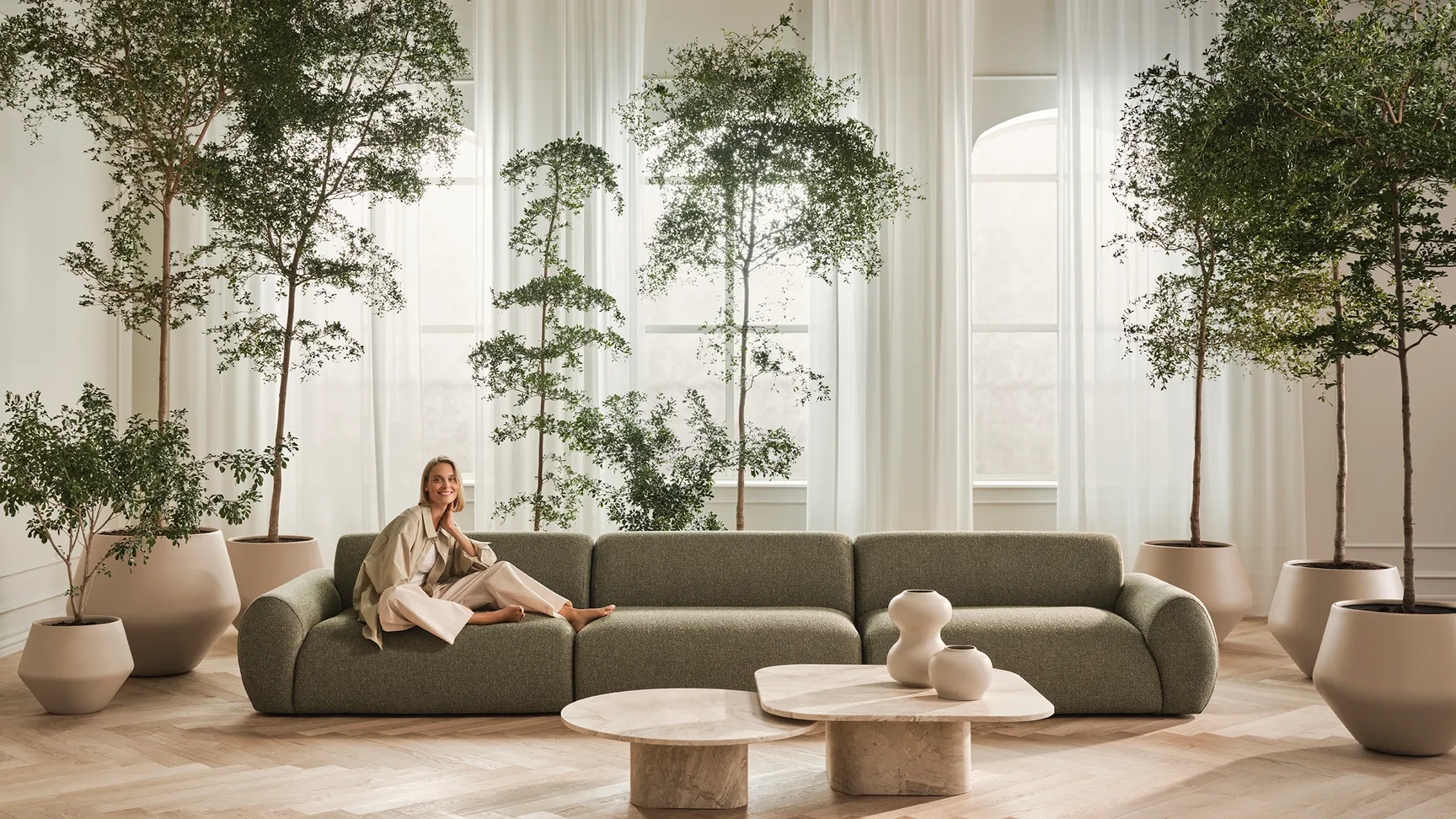 En moderne stue med en grøn sofa, en kvinde iført lyse tøj der smiler, store planter i potter, og et elegant marmorkaffebord.